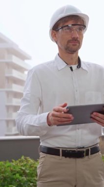 Beyaz gömlek ve kask giyen bir inşaat mühendisi ya da mimar sisli havalarda bir inşaat alanını incelerken dijital tablet kullanıyor. Mimarlık ve mühendislik