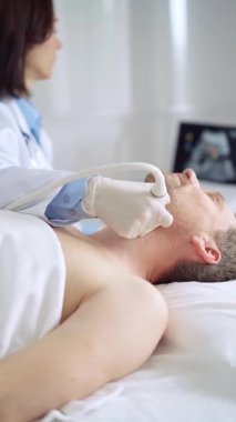 Doktor kadın, beyaz eldivenli, ultrason cihazıyla muayene masasında yatan erkek hastayı muayene ediyor.
