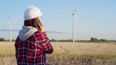 Beyaz koruyucu kask takan kadın mühendis güneş batarken rüzgar türbinleriyle dolu bir alanda akıllı bir telefonla konuşuyor. Temiz enerji ve mühendislik denetimi kavramı.