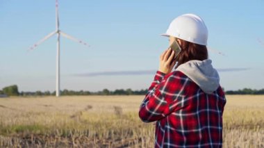 Beyaz koruyucu kask takan kadın mühendis güneş batarken rüzgar türbinleriyle dolu bir alanda akıllı bir telefonla konuşuyor. Temiz enerji ve mühendislik denetimi kavramı.