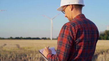 Beyaz koruyucu kask takan mühendis güneş batarken rüzgâr türbinleriyle dolu bir alanda not alıyor. Temiz enerji ve mühendislik kavramı.