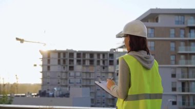 Kadın inşaat mühendisi, sabah gün doğumunda bir inşaat alanını incelerken not alıyor ve bir panoya yazıyor..