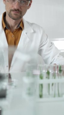 Laboratuvar önlüğü, beyaz eldiven ve koruyucu gözlük giyen bilim adamı, bir pipetten damlayan sıvıyı, içinde dikey yakın görüş olan yeşil bir bitki bulunan bir test tüpüne döküyor. Bilim ve tıp.