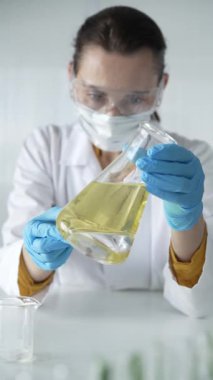 Laboratuvar önlüğü giyen, mavi koruyucu eldivenli kadın bilim adamı, laboratuvar ortamında, portre ve dikey görüntüde sarı yağlı bir Erlenmeyer şişesi tutuyor. Tıp, bilim konsepti.