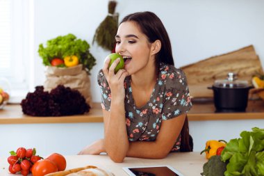 Yeşil elma tutan ve mutfakta gülümserken lezzetli bir yemek tarifini seçen çekici, genç, siyah saçlı bir kadın. Tablet pc en iyi yemek kitabıdır