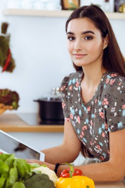 Çekici, genç, koyu saçlı bir kadın mutfakta masada otururken lezzetli bir yemeğin tarifini seçer. Tablet pc en iyi yemek kitabıdır.