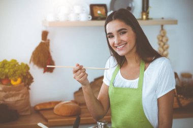Çekici, genç, siyah saçlı bir kadın, güneşli mutfakta ayakta ve gülümserken tahta kaşıkla sıcak yemek tadıyor. Yemek pişirme ve ev idaresi kavramları.