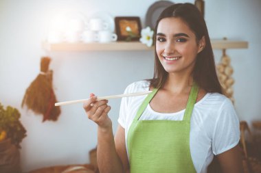 Çekici, genç, siyah saçlı bir kadın, güneşli mutfakta ayakta ve gülümserken tahta kaşıkla sıcak yemek tadıyor. Yemek pişirme ve ev idaresi kavramları.