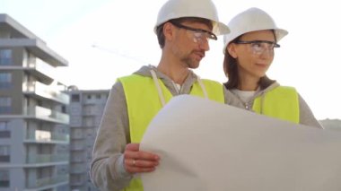 Erkek ve kadın mimarlar ya da mühendisler güvenlik kasketleri ve yelekler giyip şemaları tutuyor ve gün doğumunda inşaat alanında bir şeyler tartışıyorlar. Mimarlık ve mühendislik kavramları.