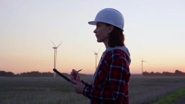 Kadın mühendis, güneş batarken rüzgâr türbinleriyle dolu bir alanda bir panoya not alıyor. Temiz enerji ve mühendislik denetimi kavramı.
