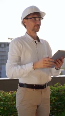 Beyaz gömlek ve kask giyen erkek inşaat mühendisi, güneş doğarken, ön ve dikey görünümde bir inşaat alanını incelerken dijital tablet kullanıyor. Mimarlık konsepti.