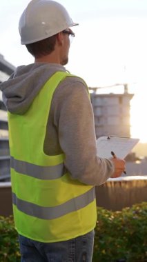 Beyaz kasklı ve güvenlik yeleği olan mühendis, sabahın erken saatlerinde dikey olarak bir inşaat alanını incelerken bir panoya not alıyor. Mimarlık ve mühendislik