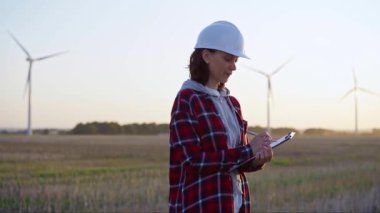 Kadın mühendis, güneş batarken rüzgâr türbinleriyle dolu bir alanda bir panoya not alıyor. Temiz enerji ve mühendislik denetimi kavramı.