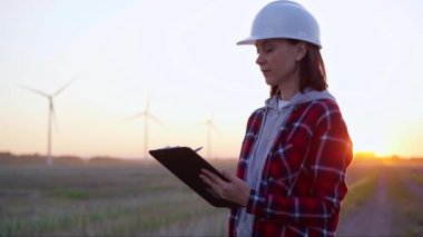 Kadın mühendis, güneş batarken rüzgâr türbinleriyle dolu bir alanda bir panoya not alıyor. Temiz enerji ve mühendislik denetimi kavramı.