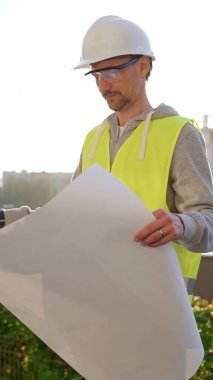 Erkek ve kadın mimarlar veya mühendisler güvenlik kasketleri ve yelekleri giyip şemaları tutuyorlar ve gün doğumunda inşaat alanında bir şey tartışıyorlar, ön dikey görüş. Mimarlık konsepti.