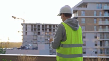 Beyaz koruyucu kasklı ve güvenlik yeleği olan adam mühendis, sabahın erken saatlerinde bir inşaat alanını incelerken panoya yazı yazıyor. Mimar konsepti.