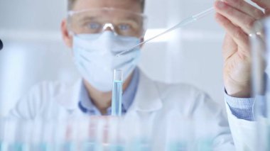 Güvenlik gözlüklü ve mavi maskeli bir adam laboratuvar tüpleriyle çalışıyor. Modern bir laboratuarda, yakın plan bir araştırmacıyı kullanıyor. Bilim ve tıp.