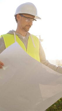 Erkek ve kadın mimarlar veya mühendisler güvenlik kasketleri ve yelekleri giyip şemaları tutuyorlar ve gün doğumunda inşaat alanında bir şey tartışıyorlar, ön dikey görüş. Mimarlık konsepti.