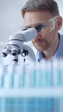 Koruyucu gözlüklü ve eldivenli bilim adamı laboratuarda mikroskop kullanıyor, araştırmacının dikey portresi. Mikrobiyoloji bilim konsepti.