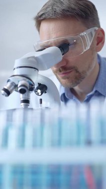 Koruyucu gözlüklü ve eldivenli bilim adamı laboratuarda mikroskop kullanıyor, araştırmacının dikey portresi. Mikrobiyoloji bilim konsepti.