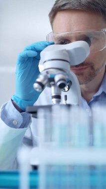 Koruyucu gözlüklü ve eldivenli bilim adamı laboratuarda mikroskop kullanıyor, araştırmacının dikey portresi. Mikrobiyoloji bilim konsepti.