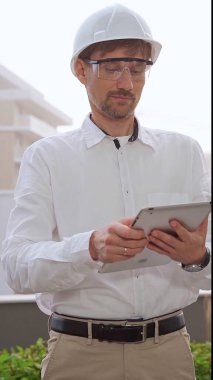 Beyaz gömlek ve kask giyen erkek mimar, sisli havada bir inşaat alanını incelerken dijital tablet kullanıyor. Mimarlık ve mühendislik konsepti.