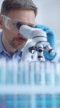 Koruyucu gözlüklü ve eldivenli bilim adamı laboratuarda mikroskop kullanıyor, araştırmacının dikey portresi. Mikrobiyoloji bilim konsepti.