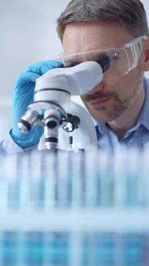 Koruyucu gözlüklü ve eldivenli bilim adamı laboratuarda mikroskop kullanıyor, araştırmacının dikey portresi. Mikrobiyoloji bilim konsepti.