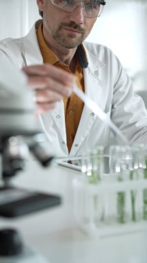 Laboratuvar önlüğü ve koruyucu gözlük takan insan bilim araştırmacısı, yeşil bir bitkiyle bir deney tüpünün içine pipetten sıvı damlatıyor. Yüzü dikey bir portreye odaklanmış. Bilim ve