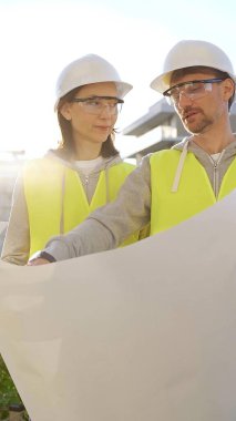 Güvenlik kaskı ve yelek giyen iki mimar ellerinde planlar tutuyorlar ve gün doğumunda inşaat alanında bir şey tartışıyorlar, dikey görüş açısı. Mimarlık mühendisliği ve takım çalışması konsepti.