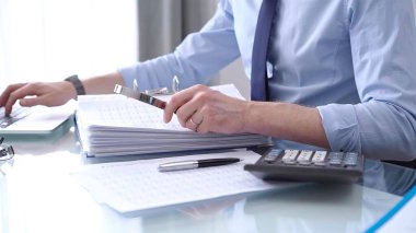 Businessman in blue short is using calculator and reviewing ring folder of financial documents with magnifying glass at desk in modern office. Audit and taxes in business concept. clipart