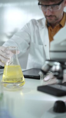 Beyaz koruyucu eldiven giyen ve gözlük takan bilim adamı bir laboratuvar dikey portresindeki Erlenmeyer şişesinin içinde sarı bir kimyasal solüsyon tutuyor. Bilim ve tıp.