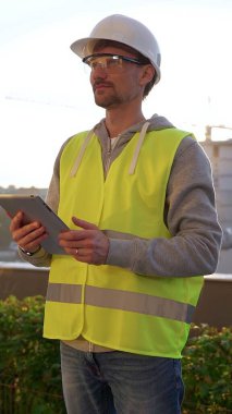 Beyaz kasklı ve güvenlik yeleği olan erkek mühendis, sabahın erken saatlerinde dikey olarak bir inşaat alanını incelerken dijital tablet üzerinde notlar alıyor. Mimarlık ve mühendislik.