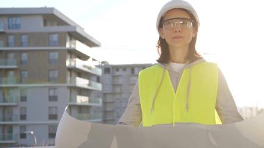 Kadın mimar ya da mühendis, güvenlik kasketi ve yelek giyiyor. Şafak vakti inşaat alanını incelerken elinde taslak var. Mimarlık konsepti.