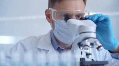 Koruyucu gözlüklü, maskeli ve eldivenli bilim adamı laboratuvarda mikroskop kullanıyor. Araştırmacı portresi. Mikrobiyoloji bilim konsepti.