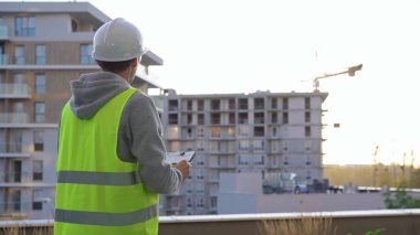 Beyaz koruyucu kasklı ve güvenlik yeleği olan adam mühendis, sabahın erken saatlerinde bir inşaat alanını incelerken panoya yazı yazıyor. Mimar konsepti.