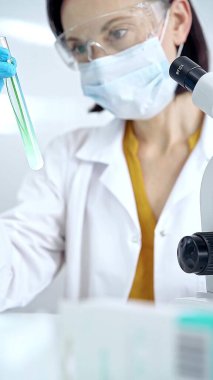 Koruyucu eldivenleri ve gözlükleri olan kadın bilim adamı yeşil sıvıyla laboratuvar tüpünü inceliyor. Mikrobiyoloji ve bilim konsepti.