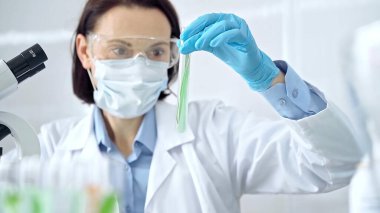 Koruyucu eldivenleri ve gözlükleri olan kadın bilim adamı yeşil sıvıyla laboratuvar tüpünü inceliyor. Mikrobiyoloji ve bilim konsepti.