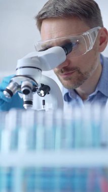 Koruyucu gözlüklü ve eldivenli bilim adamı laboratuarda mikroskop kullanıyor, araştırmacının dikey portresi. Mikrobiyoloji bilim konsepti.