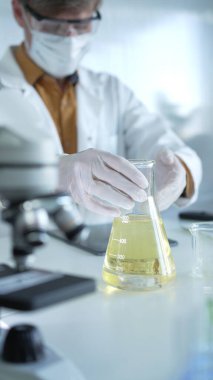 Beyaz koruyucu eldiven, maske ve gözlük takan bilim adamı araştırmacı Erlenmeyer şişesinin içinde dikey bir portre görünümünde sarı bir kimyasal solüsyon tutuyor. Bilim ve tıp.