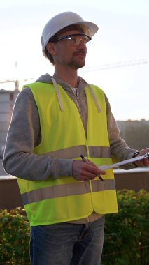 Beyaz kasklı ve güvenlik yeleği olan mühendis, sabahın erken saatlerinde dikey olarak bir inşaat alanını incelerken bir panoya notlar alıyor. Mimarlık ve mühendislik