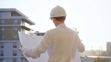 Güvenlik kaskı ve gözlük takan inşaat alanı yöneticisi, güneş doğarken veya batarken elinde planlar tutuyor ve bir inşaat alanını inceliyor. Mimarlık ve mühendislik.