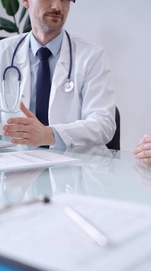 Doctor consulting patient. Male physician is gesturing above a clipboard while talking with a woman in a clinic, close up. clipart