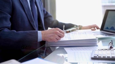 Businessman using a calculator and magnifying glass while analyzing financial documents at desk in fair modern office. Audit and taxes in business. clipart