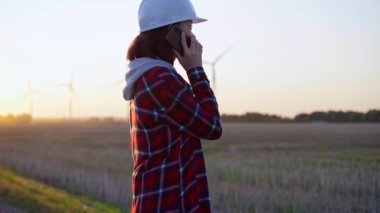 Kadın mühendis, güneş batarken rüzgâr türbinleriyle telefonda konuşuyor. Temiz enerji ve mühendislik denetimi kavramı.