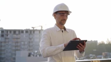 İnşaat mühendisi ya da mimar beyaz gömlekli ve başlıklı adam gün batımında bir inşaat alanını incelerken dijital tablet kullanıyor. Mimarlık ve mühendislik kavramları.