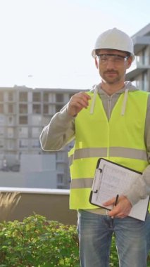 İnşaat şantiyesi mühendisi elinde bir dosya tutuyor ve arka planda gündoğumuyla birlikte yelek ve şapka giyiyor. Portre videosu
