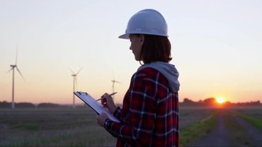 Kadın mühendis, güneş batarken rüzgâr türbinleriyle dolu bir alanda bir panoya not alıyor. Temiz enerji ve mühendislik denetimi kavramı.