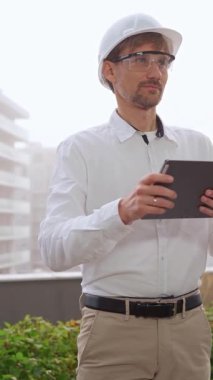 Beyaz gömlek ve kask giyen bir inşaat mühendisi ya da mimar sisli havalarda bir inşaat alanını incelerken dijital tablet kullanıyor. Mimarlık ve mühendislik