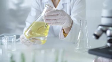 Beyaz koruyucu eldivenler ve laboratuvar önlüğü giyen tanınmayan bilim adamı laboratuvardaki masada otururken elinde sarı yağ sıvısı bulunan bir deney şişesi tutuyor. Tıp ve Bilim.